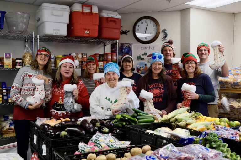 Nine people hold up holiday stockings in front of the well-stocked fresh vegetable table at the Food Share Market.