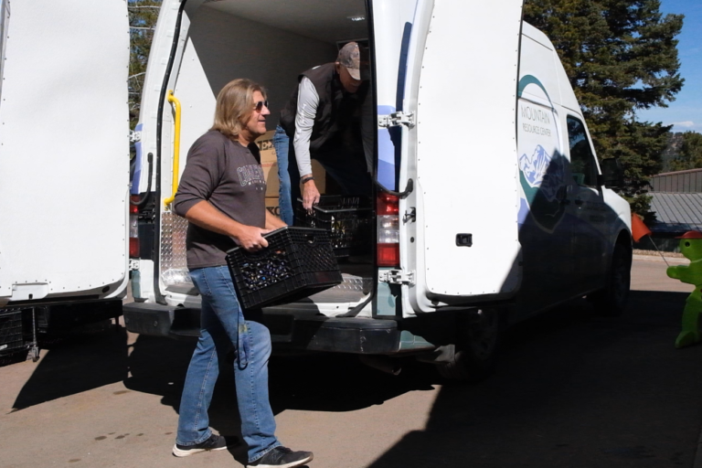 A Food Share Market volunteer wearing jeans and a dark grey shirt carries a black milk crate that has just been unloaded from a Mountain Resource Center van