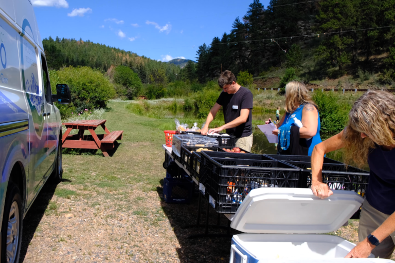 A man in tan pants and a black t-shirt, a woman in a blue dress, and a woman in tan pants and a navy blue t-shirt set up folding tables with milk crates of food on them on the right side of the image. On the left side is the Mobile Food Share Van.