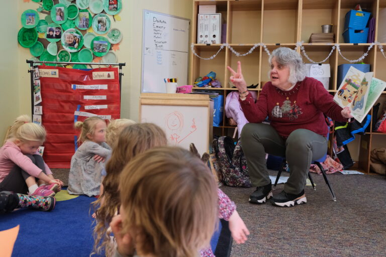 A woman in a maroon sweatshirt with holidays trees appliqued on it holds a book up for a group of children to see the picture. She is holding up two fingers on her other hand.