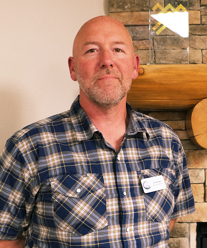 Board member Chad Atwell poses in front of a mantle in a blue and white plaid shirt.
