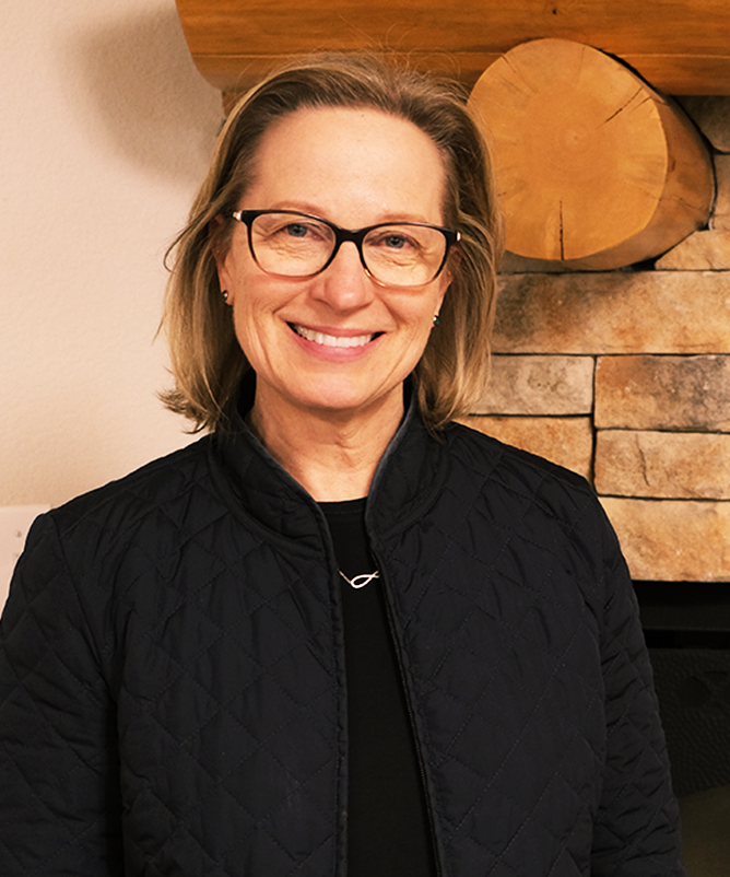 Board Member Karen Lightfoot, a woman with chin length hair and tortoiseshell glasses wearing black poses in front of a mantle