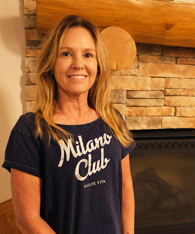 A woman with long blonde hair stands in front of a fireplace of rectangular stonework with a log mantle. She is smiling and wearing a dark navy blue, short-sleeved t-shirt that says Milano Club on the front.