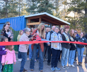 A group stands behind a long red ribbon. Two people are holding a giant pair of scissors with a red handle about to cut the ribbon.