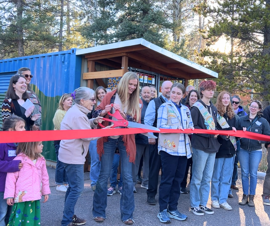 A group stands behind a long red ribbon. Two people are holding a giant pair of scissors with a red handle about to cut the ribbon.
