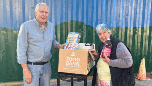 A man in a blue shirt stands on one side of a rolling cart with a box of food on top of it. Food items are showing out of the top of the box. A woman with blue hair holds up a bottle of juice on the other side of the cart.