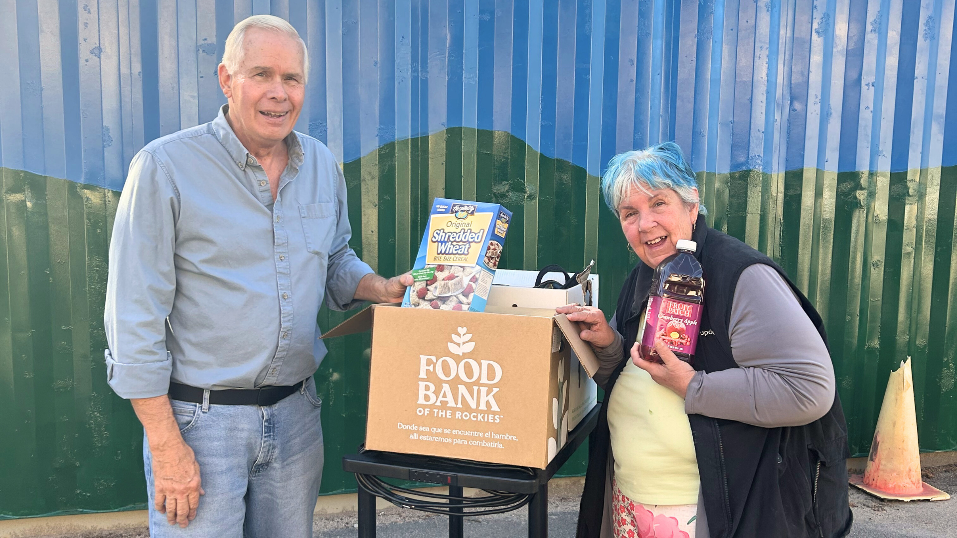 A man in a blue shirt stands on one side of a rolling cart with a box of food on top of it. Food items are showing out of the top of the box. A woman with blue hair holds up a bottle of juice on the other side of the cart.