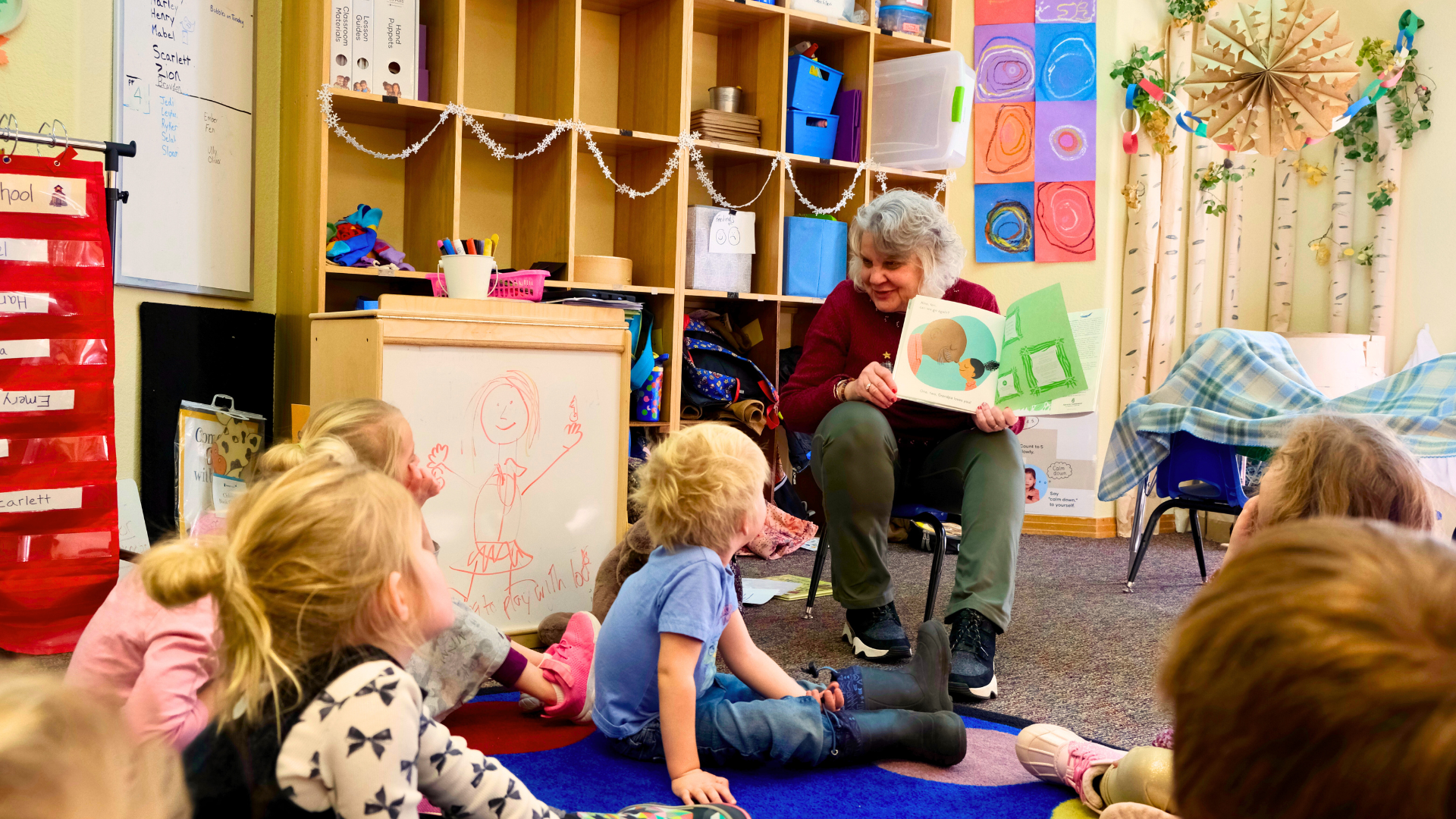 A woman sits on a small school chair showing the pages of an illustrated book to a group of children sitting on the floor.