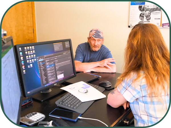 A woman in a short sleeved plaid shirt with long red hair sits at a desk with a computer, while a man in a blue t-shirt and a bandanna sits across from her.