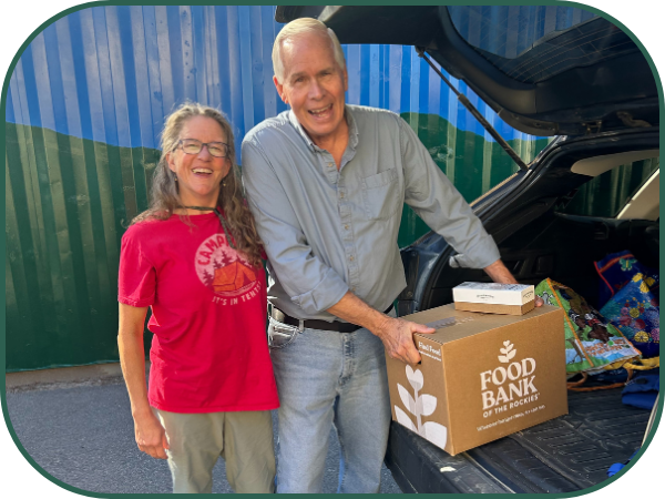 A man in blue jeans and a blue button up shirt is putting a box with the Food Bank of the Rockies logo into the back of a hatchback car. A woman wearing a red t-shirt and tan pants stands next to him posing for the picture.