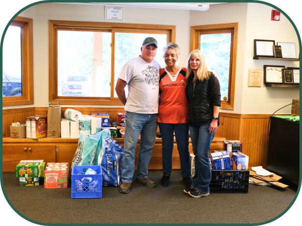 Three people stand in front of a large pile of groceries in front of some windows.