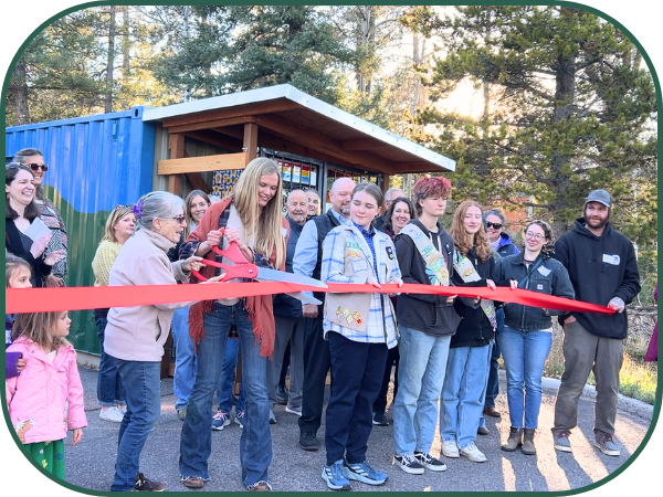 A large group of people stands in a line behind a long red ribbon. A woman with long blonde hair and and an older woman use a large novelty pair of scissors with a red handle to cut the ribbon.