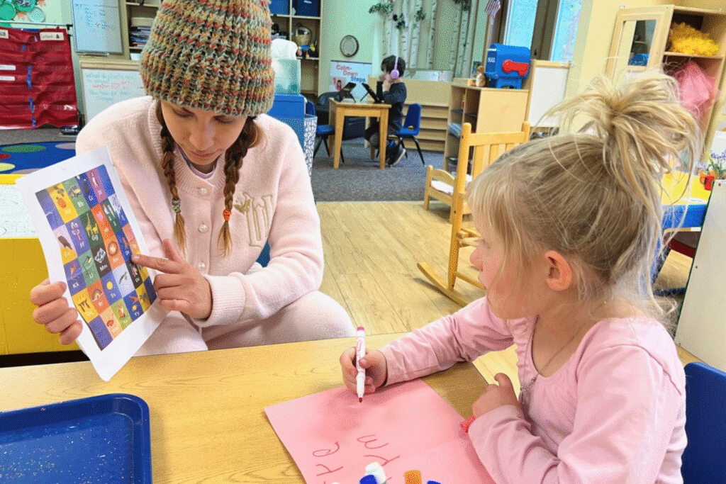 A teacher holds up an alphabet chart for a little girl in pink who is writing on a pink piece of construction paper.