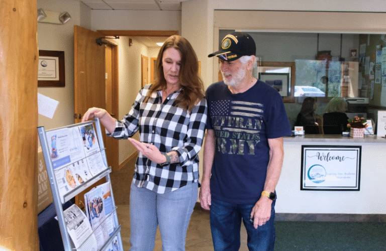 A woman in a black and white plaid shirt shows a rack of flyers to man with a white beard wearing a navy blue military t-shirt and with a veteran ball cap.