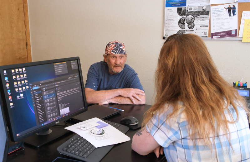 A woman with her back to the camera with a plaid shirt and long red-blonde hair sits in front of a computer assisting a man with an American flag bandana on his head and a navy blue t-shirt who sits across the desk from her facing the camera.