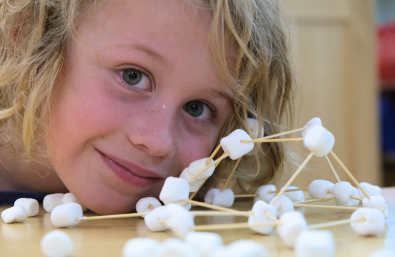 A blonde boy with long slightly curly hair poses with his face near to a structure he has built from small marshmallows and toothpicks.