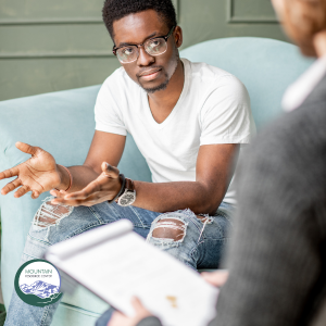 A man in fashionably ripped jeans and a white t-shirt is facing the camera. He has his hands out as if he is explaining something. On the right side of the image, an out of focus person holds a notepad.
