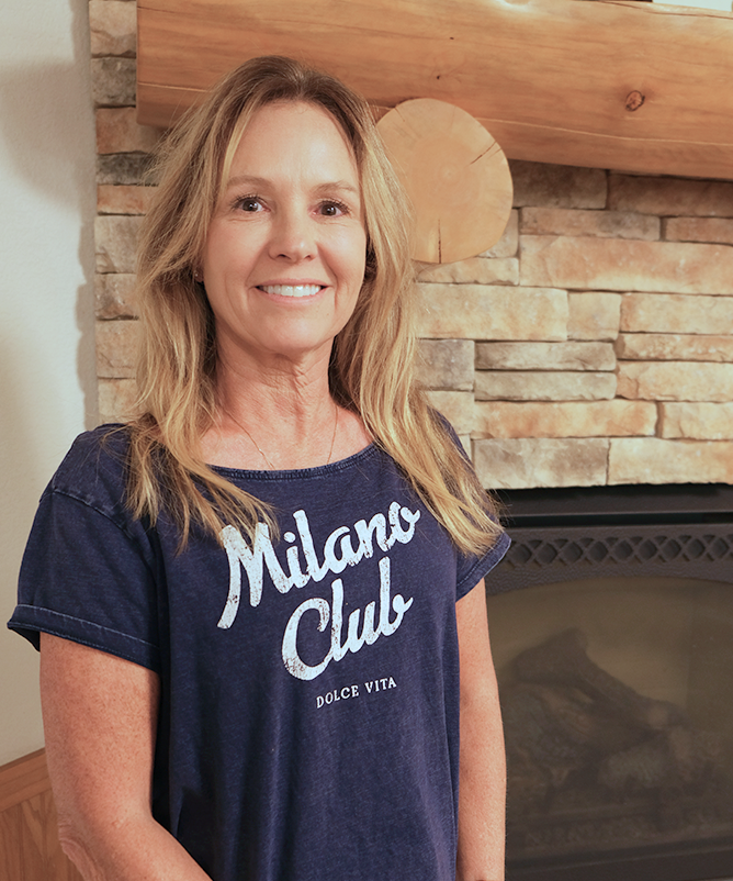 A woman with long blonde hair stands in front of a fireplace of rectangular stonework with a log mantle. She is smiling and wearing a dark navy blue, short-sleeved t-shirt that says Milano Club on the front.