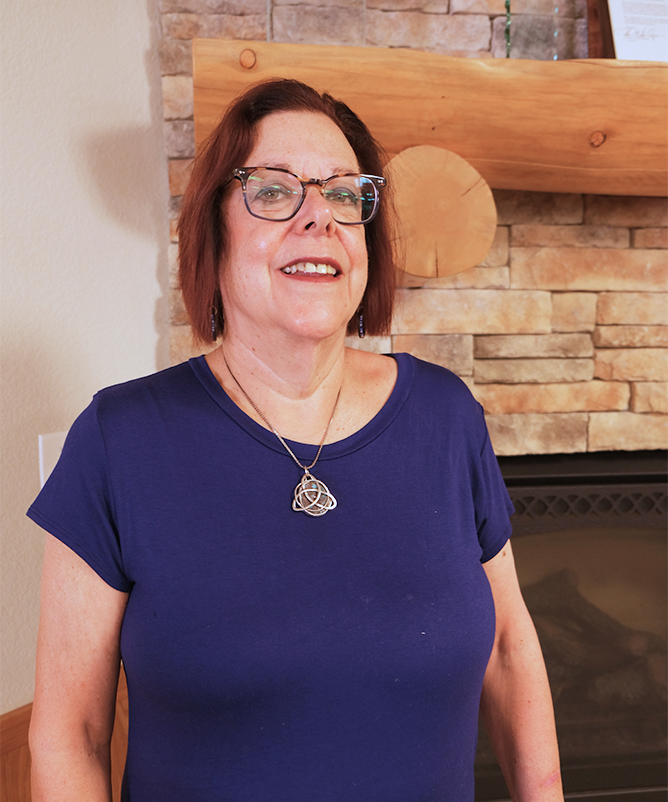 A woman with dark red hair, glasses, and dark lipstick stands in front of a fireplace with rectangular stonework and a log mantle. She is wearing a short-sleeved, dark navy blue t-shirt with a scoop neck, and a pendant necklace.