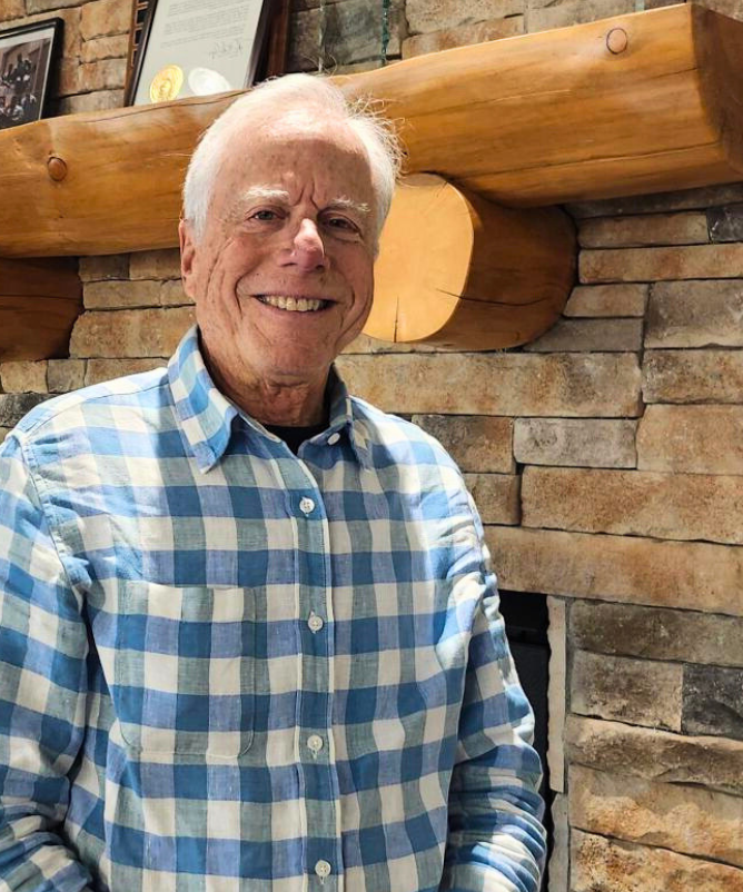 A smiling man with white hear stands in front of a fireplace at MRC wearing a blue checked button up shirt.
