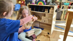 Two preschoolers in blue shirts are pictured. One has her back to the camera and is partially out of frame on the left. She is handing the other student a stack of square blocks to put into a dedicated block shelf with places for each kind of block.