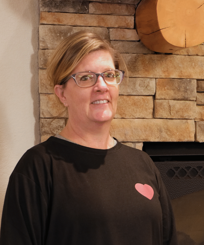 A blonde woman with glasses stands in front of a brick fireplace at MRC. She is wearing a long-sleeved black t-shirt with a small heart patch on the upper left.