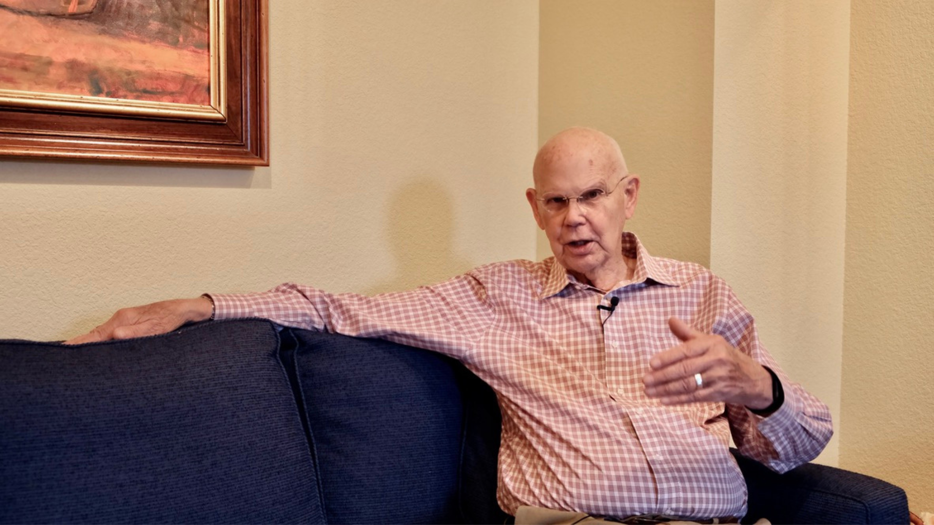 An older man in a red and white graph check button-up shirt with long sleeves sits on a blue couch with his arm outstretched along the back. His other hand is gesticulating as he tells a story.