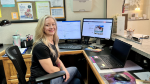 A long-haired blonde woman in a fitted black t-shirt sits behind the front desk at Mountain Resource Center. Two computer monitors are visible behind her.