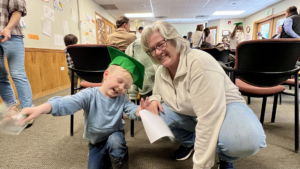 A grandma in an oatmeal colored sweater crouches with her grandson, who is wearing a blue sweatshirt and a bright green mortarboard hat. His eyes are closed and he has a delighted smile on his face. One hand is outstretched in a joyful gesture, and the other, closest to his grandma, holds his diploma.