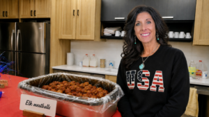 A woman with long dark hair wearing a black sweatshirt with USA in a US flag pattern stands next to a warming pan of elk meatballs.