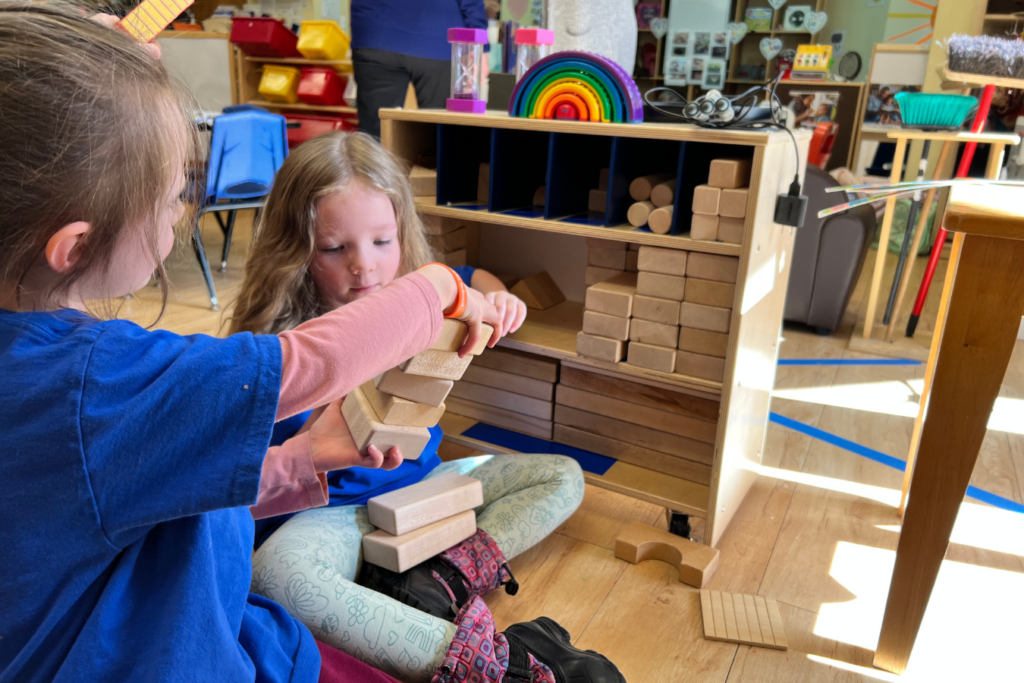 Two young children in blue t-shirts are putting blocks away in our preschool classroom. One girl is handing the other a stack of blocks to go onto the shelf.