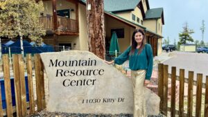 A dark-haired woman in a teal blouse and gold trousers, Sarah Kinzer, stands next to the flagstone Mountain Resource Center sign in front of the MRC building.