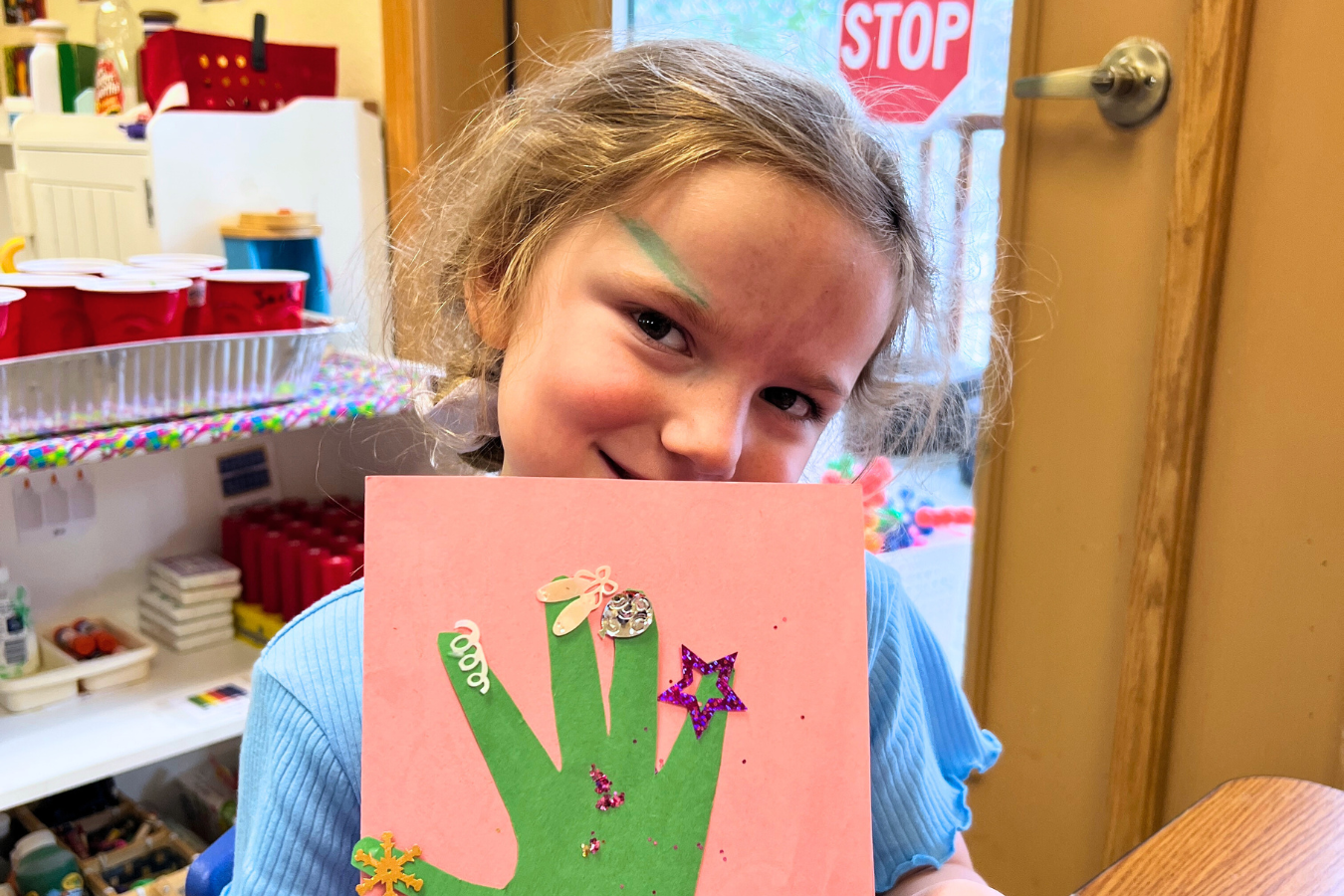 A little girl in a blue t-shirt holds up a pink rectangle of construction paper. A green handprint, also made of construction paper, is glued in the center. The tips of the fingers are decorated with cutout shapes.