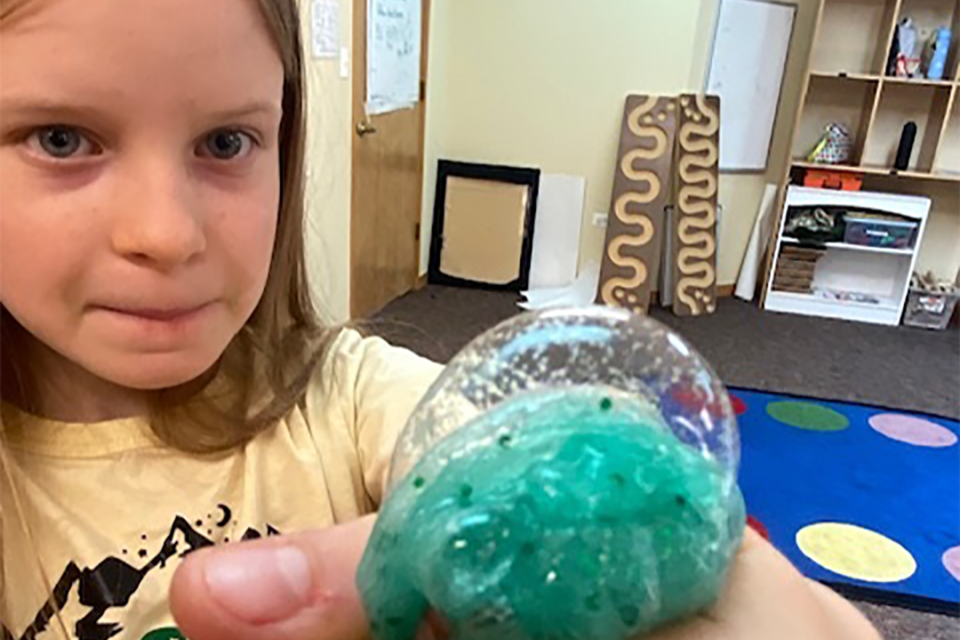 A girl in a yellow shirt stares intently at a bubble of green slime in her outstretched hand.
