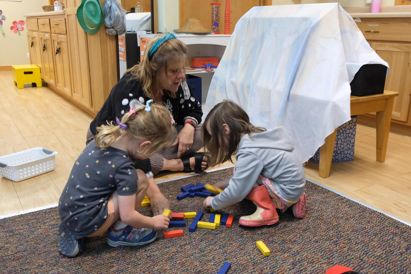 Preschool director Carla sits on the floor with two young girls teaching math with colorful lengths of wood.