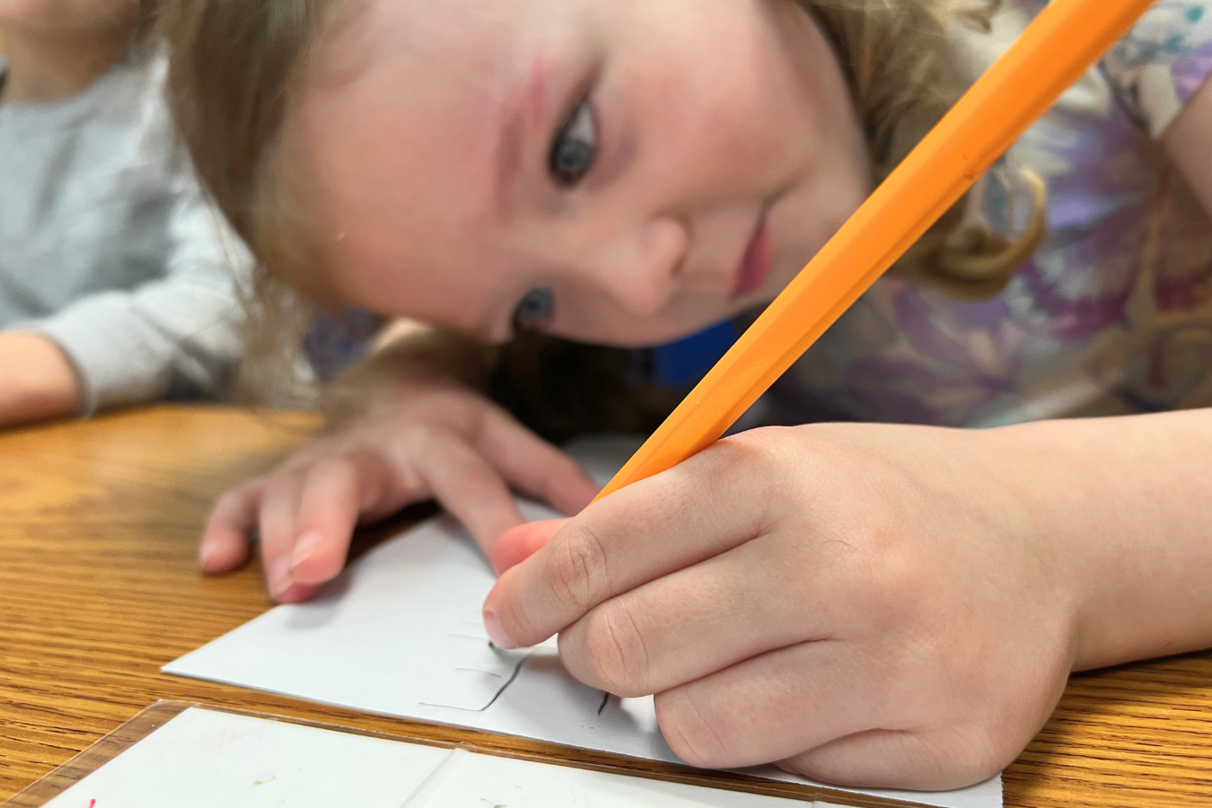 An out of focus preschool student is bent low over a table in the background. In focus in the foreground is a close up of her hand, pencil, and paper as she carefully makes letters.